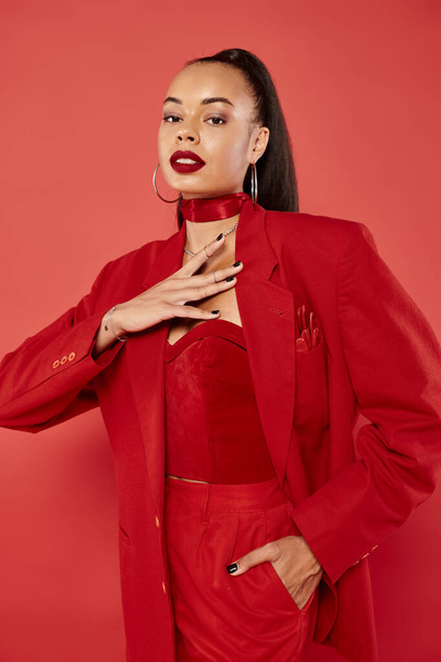beautiful african american woman with ponytail posing in suit with hand near chest on red background - Photo, Image