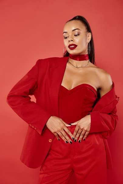 attractive african american woman with ponytail and straight hair posing in suit on red background - Photo, Image