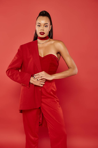 elegant african american woman with ponytail posing in vibrant formal wear on red backdrop - Photo, Image