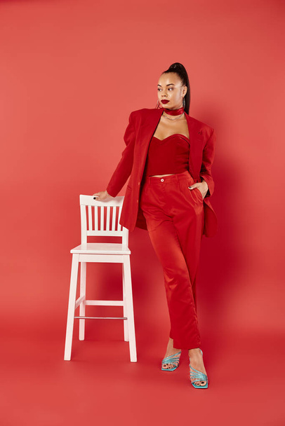 longitud completa de elegante mujer afroamericana posando en traje rojo con la mano en el bolsillo cerca de la silla - Foto, Imagen
