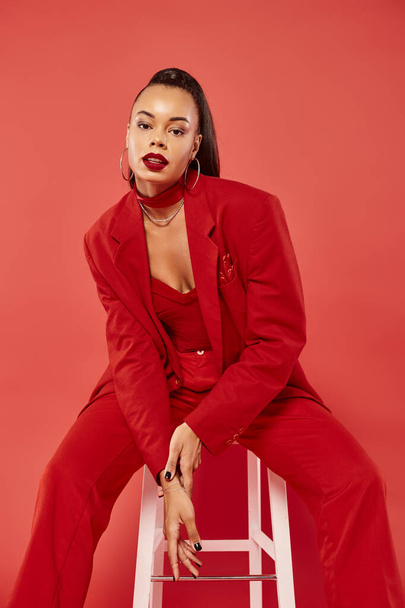 beautiful brunette african american woman in suit sitting on white chair on red background - Foto, imagen