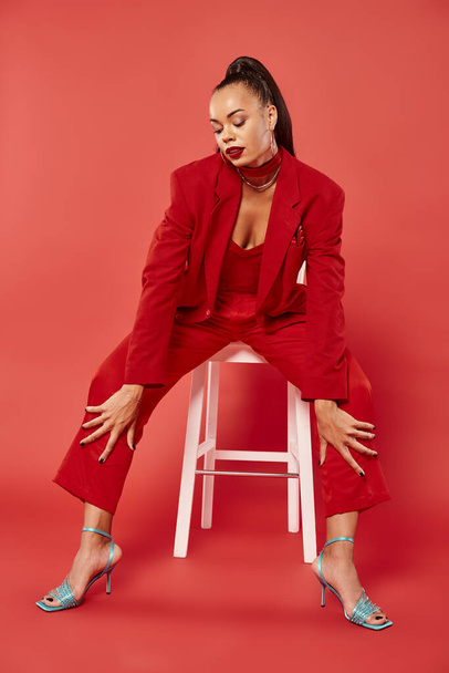 volledige lengte van elegante Afrikaanse Amerikaanse vrouw in pak en hoge hakken zitten op witte stoel op rood - Foto, afbeelding