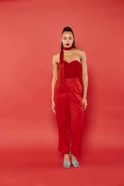 longitud completa de atractivo modelo afroamericano en top sin tirantes y pantalones posando sobre fondo rojo - Foto, Imagen
