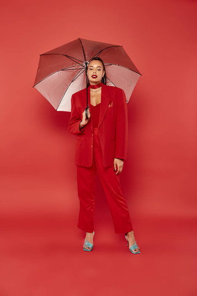volledige lengte, brunette Afrikaans amerikaans model in rode blazer en broek staande onder paraplu - Foto, afbeelding