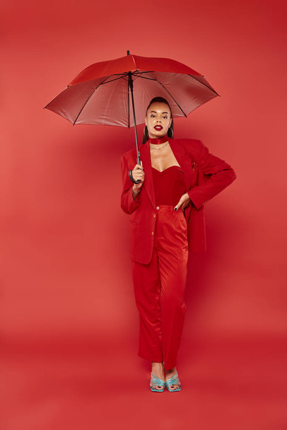 mujer afroamericana bonita en pantalones rojos y chaqueta de pie bajo paraguas, mano en la cadera - Foto, Imagen