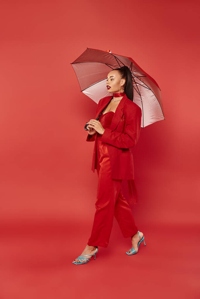 csinos afro-amerikai nő blézerben és nadrág séta esernyő alatt piros háttér - Fotó, kép