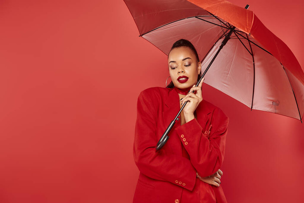 attractive young african american woman in blazer and pants standing under umbrella on red backdrop - Photo, Image
