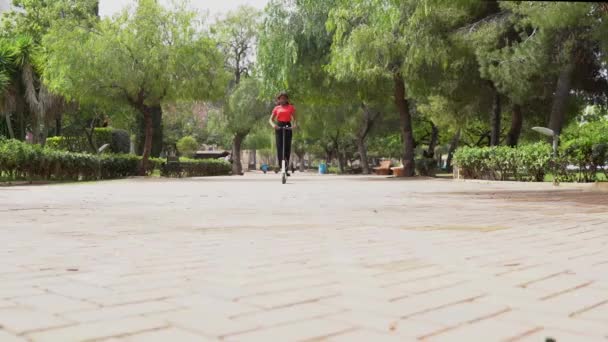 Reaaliaikainen Afrikkalainen amerikkalainen nainen ratsastus sähkö skootteri päällystetty kävelytie kadulla vihreitä puita kaupungin kesäpäivänä - Materiaali, video