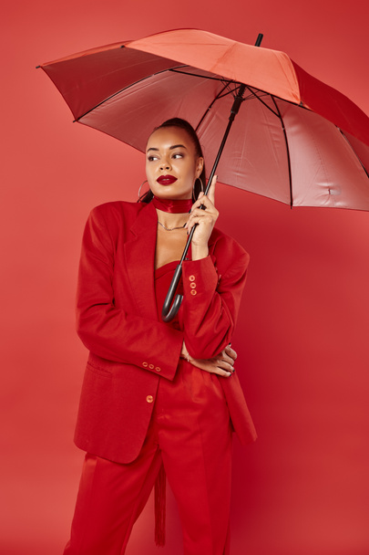 attractive young african american woman in jacket and pants standing under umbrella on red backdrop - Photo, Image
