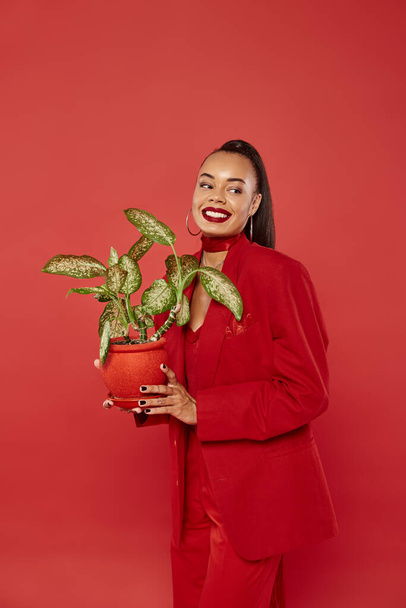 gelukkig jong Afrikaans amerikaans vrouw in rood pak jas en broek staan met potted groen plant - Foto, afbeelding