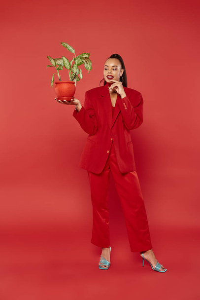 longitud completa, mujer afroamericana feliz en ropa formal roja de pie con maceta planta verde - Foto, Imagen
