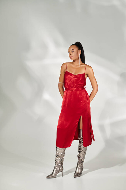 Full length, όμορφη Αφροαμερικανή γυναίκα με κόκκινο φόρεμα και ασημένιες μπότες που ποζάρουν σε γκρι φόντο - Φωτογραφία, εικόνα