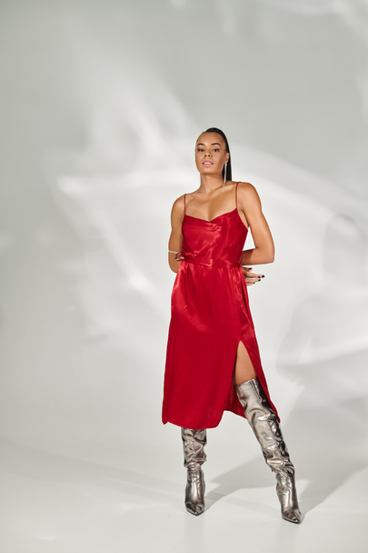 full length, confident african american woman in red dress and silver boots posing on grey backdrop - Photo, Image