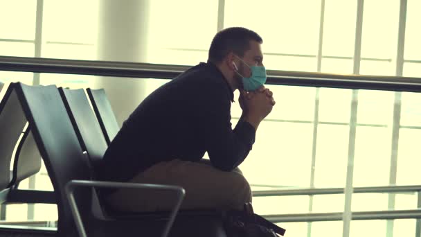 jonge man met een beschermend gezichtsmasker zittend in de luchthaventerminal, mobiele telefoon vasthoudend en wachtend op de vlucht. - Video