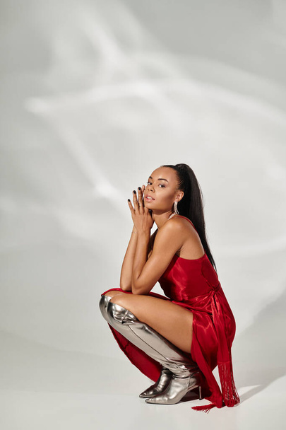 pretty african american woman in red midi dress and silver boots sitting on mirrored grey backdrop - Photo, Image