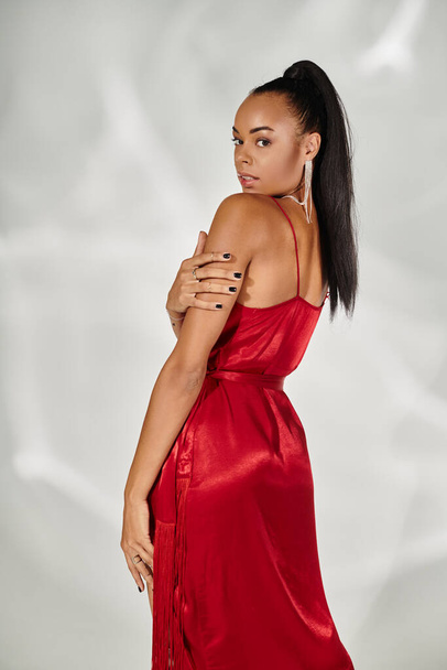 alluring african american woman in red dress looking at camera and posing on mirrored grey backdrop - Photo, Image