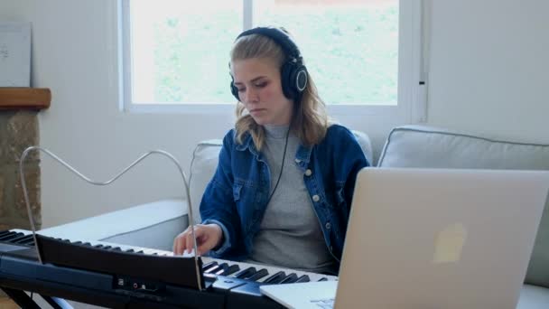 Fiatal nő alkalmi ruhát fejhallgatóval videó bemutató a laptopon, és játszik elektronikus zongora, miközben ül a hangulatos szobában kandallóval a modern lakás - Felvétel, videó