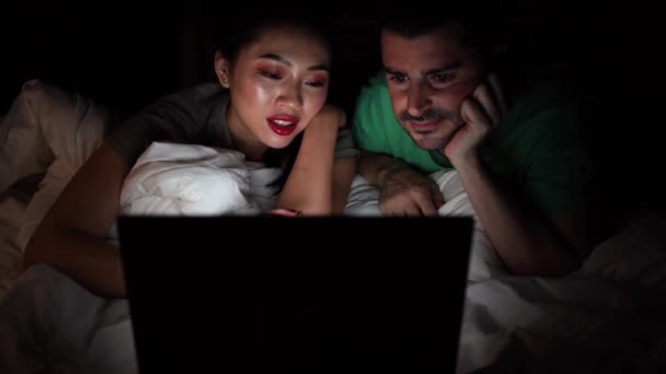 Азиатка лежит на кровати рядом с контентом бойфренд в повседневной одежде во время просмотра фильма на ноутбуке дома. - Кадры, видео