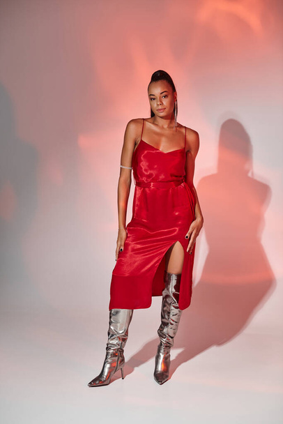 attractive african american woman in midi red dress and silver boots on grey backdrop with lighting - Photo, Image