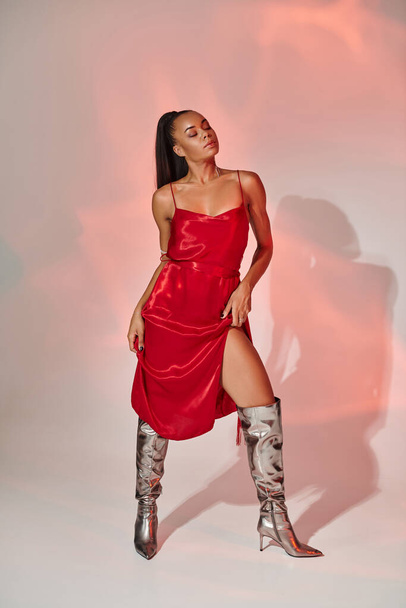 full length, african american woman in red midi dress and silver boots posing on grey with lighting - Photo, Image
