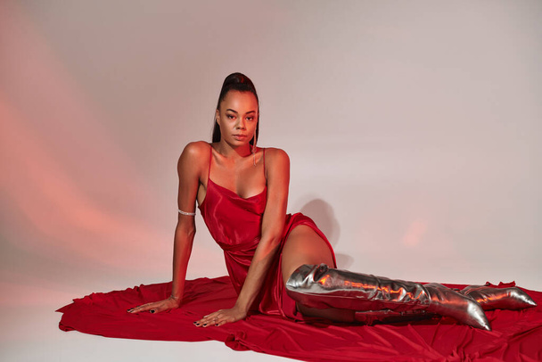 attractive african american woman in red midi dress and silver boots sitting on grey with lighting - Photo, Image