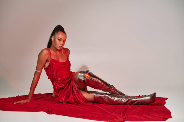 attractive african american woman in red midi dress and silver boots sitting on grey with lighting - Photo, Image