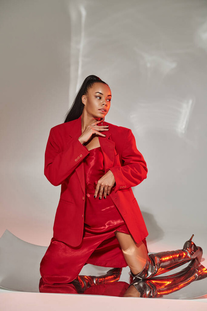 young african american woman in red blazer, dress and silver boots posing on mirrored surface - Photo, Image