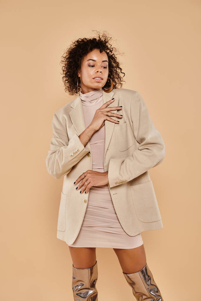 brunette african american woman with curly hair posing in autumnal blazer and dress on beige - Photo, Image