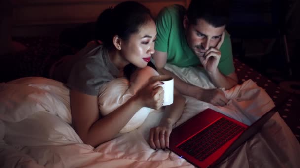 Asiática mujer acostada en una cama cerca de contenido novio en ropa casual mientras mira una película en un ordenador portátil en casa. - Metraje, vídeo