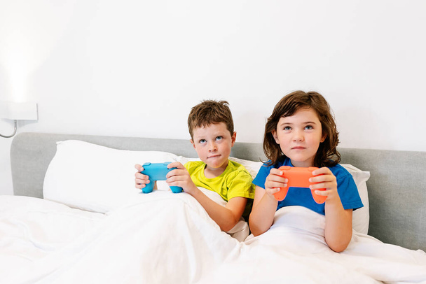 Hermanos enfocados con gamepads jugando interesante videojuego mientras están sentados juntos en una cómoda cama bajo una manta en el dormitorio en casa - Foto, Imagen
