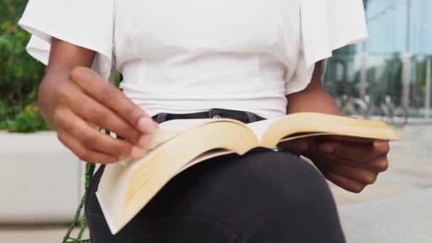 Cropped unrecognizable young African American female in stylish clothes with long braided hair flipping pages and reading story in book while sitting on bench on windy day in city - Footage, Video