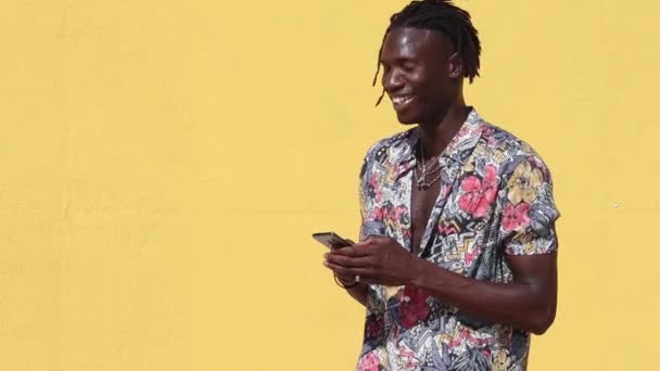 Cheerful young hipster African American male with dreadlocks wearing trendy summer shirt with floral print and jeans using mobile phone against yellow background - Footage, Video