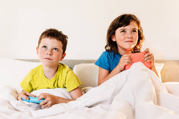 Hermanos enfocados con gamepads jugando interesante videojuego mientras están sentados juntos en una cómoda cama bajo una manta en el dormitorio en casa - Foto, imagen