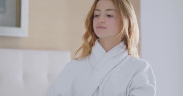 Młoda kobieta millennial w miękkim białym szlafroku zdjąć okulary i przesuwając jej rude włosy w jasnej sypialni - Materiał filmowy, wideo