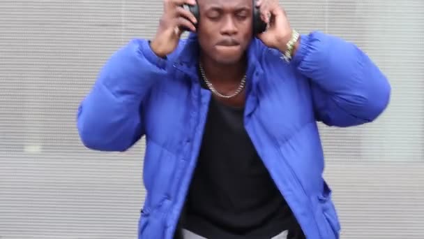 Hombre étnico calmo que usa un elegante atuendo hipster parado en un edificio de hormigón en la calle con auriculares y disfrutando de las canciones - Metraje, vídeo
