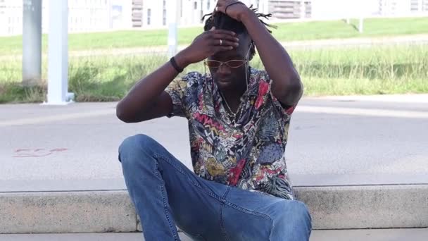 Baixo ângulo de homem afro-americano sério em camisa colorida e jeans sentado em degraus de pedra e fazendo um rabo de cavalo na rua urbana - Filmagem, Vídeo