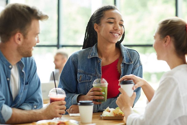 Smiling attractive young Black woman in denim jacket standing at table and enjoying time with friends - Foto, Imagem