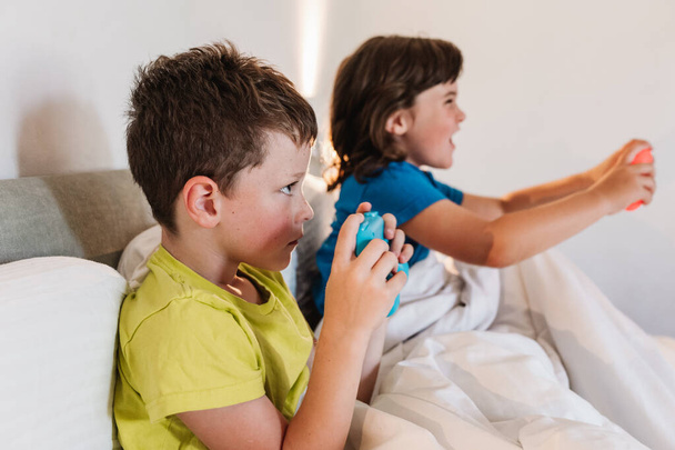 Vista lateral de los hermanos enfocados con gamepads jugando interesante videojuego mientras están sentados juntos en una cómoda cama bajo una manta en el dormitorio en casa - Foto, imagen
