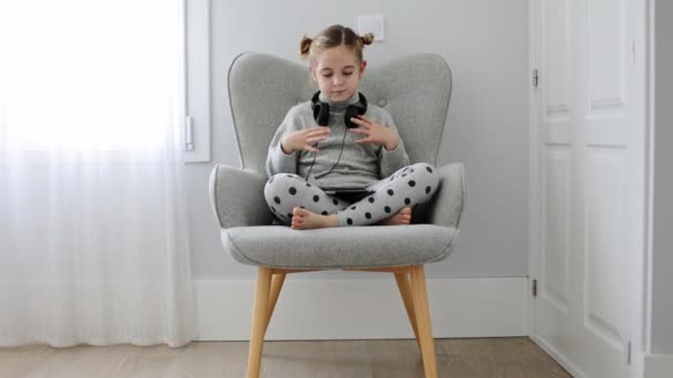 Full body of barefoot girl sitting with crossed legs in armchair and listening to songs from tablet in headphones - Footage, Video