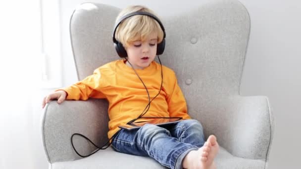 Full body of cute barefoot little boy in headphones sitting in armchair and watching cartoon on digital tablet at home - Footage, Video
