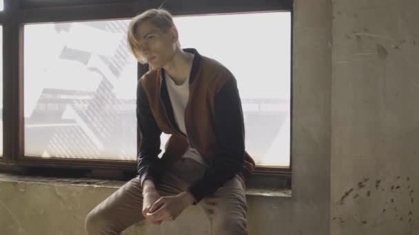 Atrakcyjny młody blondyn siedzący na parapecie w pobliżu dużych okien w zakurzonym pokoju z porysowanymi ścianami i odwracając wzrok - Materiał filmowy, wideo