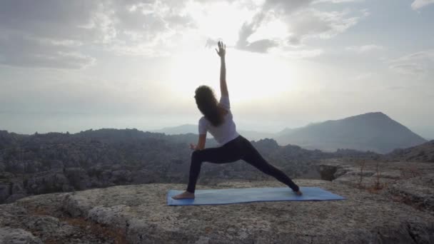 Vista laterale a basso angolo della femmina flessibile in piedi nella posizione Lord of the Dance contro il cielo nuvoloso del tramonto mentre pratica yoga in cima alla montagna rocciosa - Filmati, video