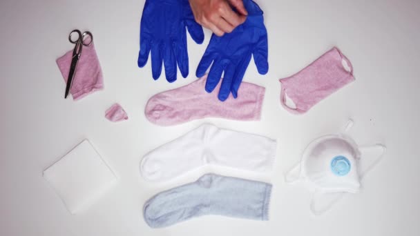 Зверху безликі люди в синіх стерильних рукавичках кладуть білу серветку всередину бавовняного носка, роблячи маску для обличчя вдома під час COVID 19
 - Кадри, відео