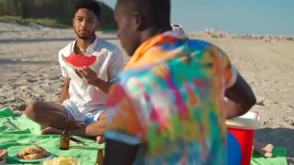 Szczęśliwy wielonarodowy mężczyzna i kobiety w luźnych ubraniach uśmiechający się i jedzący owoce siedząc na kocu i słuchając czarnego przyjaciela grającego na gitarze akustycznej w pobliżu morza w letni weekend na plaży - Materiał filmowy, wideo