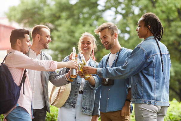 Group of cheerful young interracial people standing in city park and clinking bottles while drinking to friendship - Φωτογραφία, εικόνα