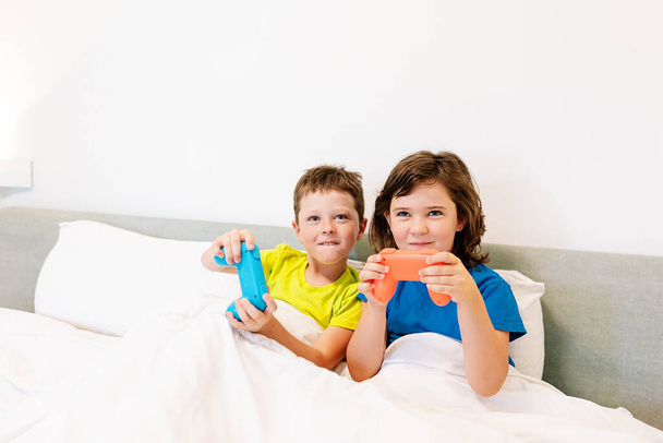 Hermanos contentos con gamepads jugando interesante videojuego mientras están sentados juntos en una cómoda cama bajo una manta en el dormitorio en casa - Foto, Imagen