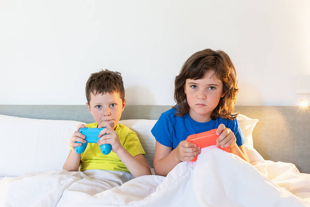 Zaostření sourozenci s gamepady hrát zajímavé videohry, zatímco sedí spolu v pohodlné posteli pod dekou v ložnici doma - Fotografie, Obrázek