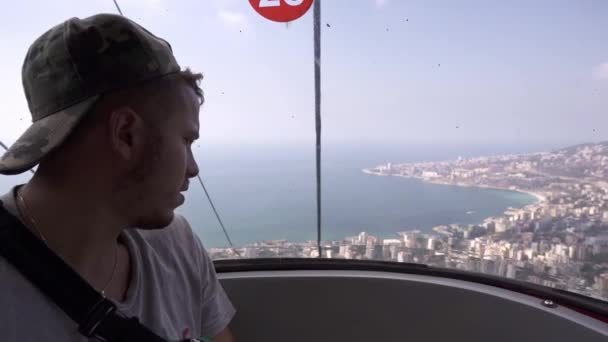 Молодой путешественник в повседневной одежде сидит в кабине гондолы лифта и любуется удивительными пейзажами прибрежного города Джуние в солнечный день во время поездки в Ливан - Кадры, видео