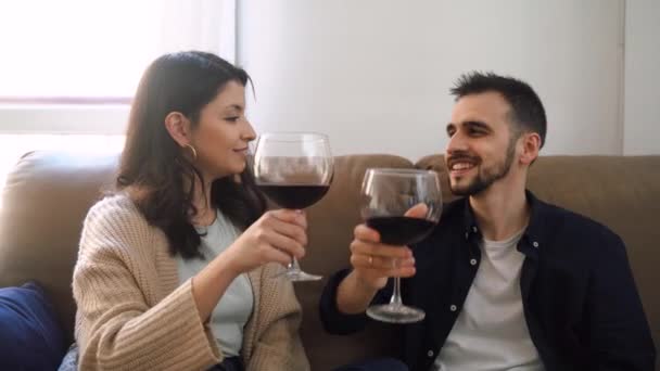Χαλαρωμένο ζευγάρι με κόκκινο κρασί σε ποτήρια που κάθονται στον καναπέ και κοιτάζονται ενώ περνούν το Σαββατοκύριακο μαζί - Πλάνα, βίντεο