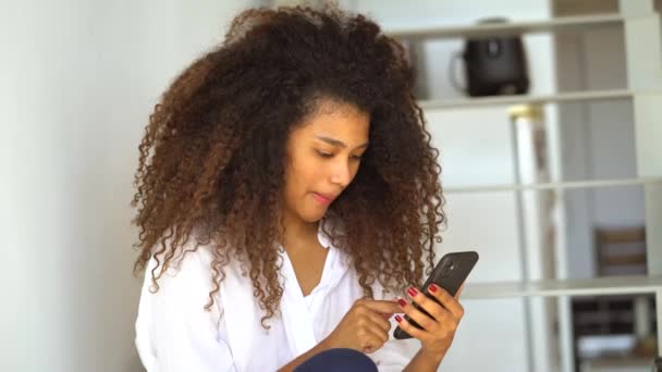Young African American female in casual outfit surfing internet on mobile phone while resting in living room at home - Footage, Video